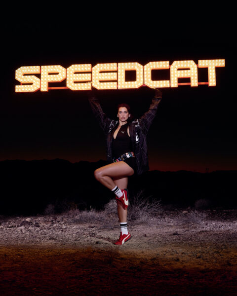 Piosenkarka Dua Lipa ubrana w czarny top oraz marynarkę i czerwone buty Puma Speedcat oraz białe skarpetki pozuje trzymając w rękach pomarańczowy napis Speedcat.
