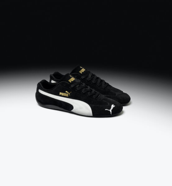 Czarne buty Puma Speedcat, które mają białe akcenty oraz złoty napis, położone są na biało-czarnym tle.