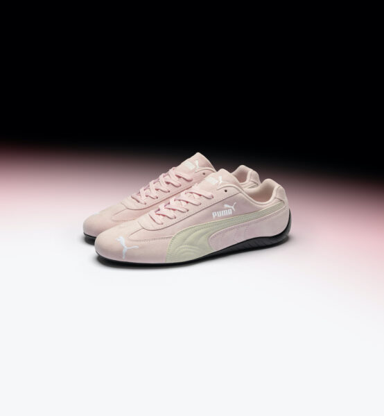 Różowe buty Puma Speedcat, które mają białe akcenty oraz złoty napis, położone są na różowo-czarnym tle.