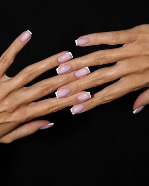 Splecione dłonie na czarnym tle. Dłonie mają długie, smukłe palce oraz kwadratowe paznokcie pomalowane w stylu french manicure.