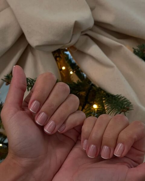 Paznokcie w stylu french manicure na krótkich paznokciach o kształcie kwadratu. Dłonie z manicure znajdują się na tle choinki ozdobionej dużą, beżową kokardą.
