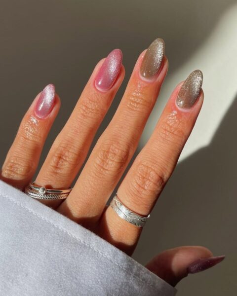 Dłoń z długimi, owalnymi paznokciami, które zdobi magnetyczny manicure w kolorze złotym i różowym. Na palcu serdecznym i wskazującym widać pierścionki. Widać też fragment rękawa białej koszuli.