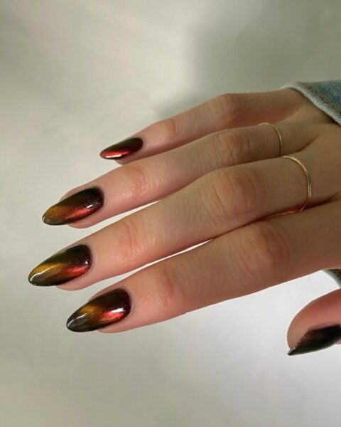 Manicure magnetyczny w kolorze czarnym z czerwonym poblaskiem na długich, migdałowych paznokciach. Na palcu środkowym i serdecznym znajdują się delikatne, złote obrączki.