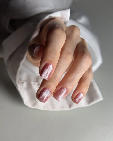 Zbliżenie na dłoń z długimi, kwadratowymi paznokciami z manicure magnetycznym w kolorze jasnoróżowym. Widać w tle rękaw białej koszuli. Dłoń pozuje na białym tle.