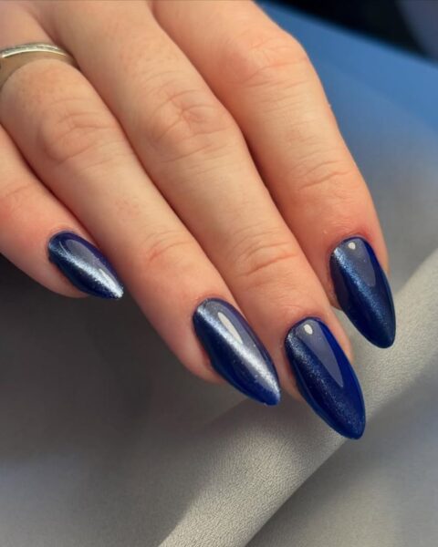 Manicure magnetyczny w kolorze granatowym na długich, migdałowych paznokciach. Dłoń pozuje na tle szarego materiału. Na palcu serdecznym widać pierścionek z brylantem.