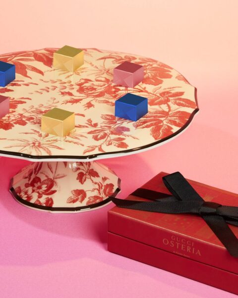 Porcelanowa patera marki Gucci ozdobiona czerwonym floralnym wzorem na jasnym tle, ustawiona na różowym tle. Na paterze znajdują się kolorowe pudełeczka w kształcie kostek. Obok leży eleganckie czerwone pudełko z napisem "Gucci Osteria", przewiązane czarną wstążką.