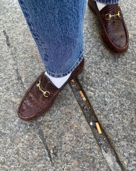 Zdjęcie przedstawia fragment stylizacji, na którym widoczne są jeansowe spodnie rurki oraz eleganckie, brązowe loafersy z fakturą imitującą skórę krokodyla, ozdobione złotymi detalami. Buty zostały zestawione z białymi skarpetkami, co nadaje stylizacji casualowo-elegancki charakter. Tło to chodnik z wyraźnymi śladami użytkowania, w tym niedopałkami papierosów, co tworzy kontrast między luksusowymi elementami ubioru a surowym, miejskim otoczeniem.