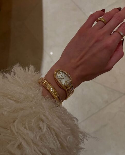 Luksusowe zegarki damskie w eleganckiej stylizacji. Na zdjęciu widoczna jest kobieca dłoń ozdobiona złotym zegarkiem o biżuteryjnym designie, z opływową tarczą oraz diamentowymi zdobieniami. Obok zegarka znajduje się złota bransoleta, a palce zdobią subtelne pierścionki z perłami i kamieniami szlachetnymi. Stylizacja uzupełniona jest kremowym futrzanym płaszczem, co podkreśla ekskluzywny charakter dodatków. Idealny przykład ponadczasowej elegancji w kategorii luksusowe zegarki damskie.