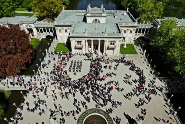 Tłum ludzi zgromadzony przed Pałacem na Wyspie w Łazienkach Królewskich w Warszawie, widziany z lotu ptaka podczas słonecznego dnia. Na dziedzińcu odbywa się wydarzenie plenerowe, widoczne są ustawione krzesła i scena, wokół której gromadzą się uczestnicy.