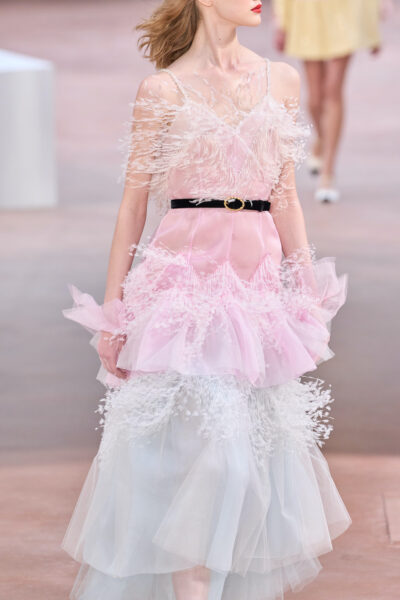 Modelka prezentuje romantyczną suknię haute couture od Chanel w pastelowych odcieniach różu i błękitu podczas pokazu na sezon lato 2025. Kreacja wykonana z warstw tiulu i ozdobiona subtelnymi piórkami emanuje lekkością i bajkowym wdziękiem. Czarno-złoty pasek dodaje kontrastu i elegancji. Stylizacja doskonale oddaje trend na eteryczne, cukierkowe pastele oraz zmysłowe tekstury inspirowane naturą, które będą dominować w letnich kolekcjach 2025.