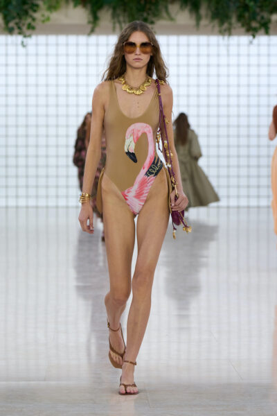 Modelka prezentuje jeden z najbardziej charakterystycznych looków z kolekcji trendy bikini 2025 — jednoczęściowy, beżowy kostium kąpielowy z dużym nadrukiem różowego flaminga i podpisem "Chloé". Stylizację uzupełniają złote akcesoria, duże okulary przeciwsłoneczne i sandały, a całość wpisuje się w egzotyczny, luksusowy klimat pokazu mody na tle roślin i białej kratki.