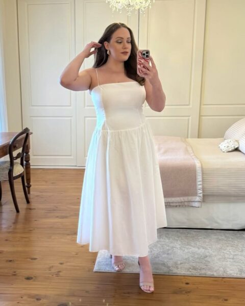 Zdjęcie kobiety plus size. Ma ciemne, rozpuszczone włosy do ramion. Ubrana jest w białą sukienkę rozkloszowaną i na ramiączkach. Ma do tego beżowe sandałki na obcasie. Pozuje w sypialni i robi sobie selfie w sukience.