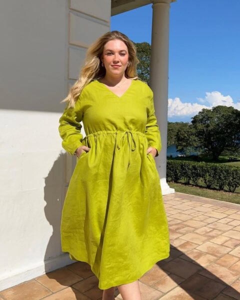 Zdjęcie kobiety plus size z blond włosami rozpuszczonymi na ramiona. Ma na sobie sukienkę dla niskiej osoby z brzuszkiem, czyli kopertową z wiązaniem w pasie, w kolorze zielonym Ma też kieszenie, w których trzyma ręce. Pozuje na tarasie.