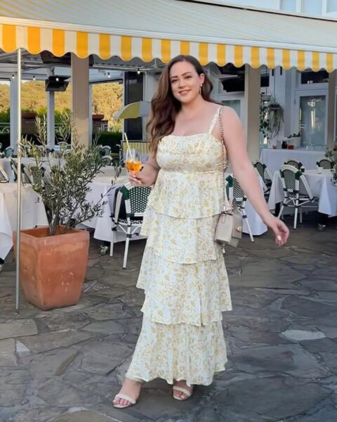 Zdjęcie kobiety plus size z ciemnymi włosami. Ma na sobie kremową, kwiecistą sukienkę na ramiączkach o fasonie empire i z falbankami na dole. Do tego ma beżowe sandałki na obcasie oraz małą, kremową torebkę na ramieniu. Pozuje na tle restauracji w modnym kurorcie.