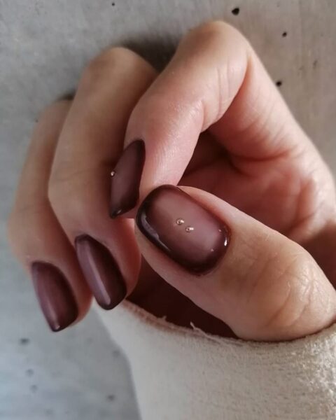 Zdjęcie dłoni z średniej długości, kwadratowymi paznokciami w kolorze brązowym z efektem aura nails. Do tego na środku płytki umieszczono drobne, złote aplikacje. Dłoń pozuje na tle szarej ściany.