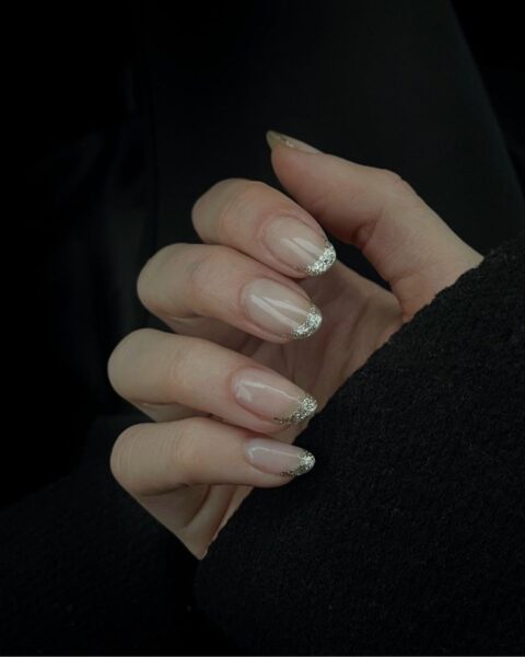 Zdjęcie dłoni ze zbliżeniem na średniej długości paznokcie o owalnym kształcie z brokatowym french manicure w kolorze srebrnym. Dłoń pozuje na czarnym tle.