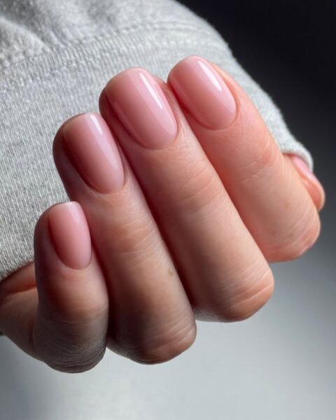 Zdjęcie dłoni ze zbliżeniem na manicure dla paznokci średniej długości. Mają kwadratowy kształt i są ozdobione stylizacją glazed donut nails w kolorze różowym z mocnym połyskiem. Dłoń pozuje na tle białego blatu.