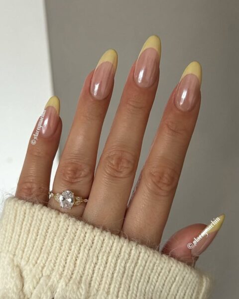 Zdjęcie dłoni z długimi, migdałowymi paznokciami z french manicure w kolorze żółtym. Na palcu znajduje się złoty pierścionek z brylantem. Widać też fragment rękawa swetra z kolorze maślanej żółci.