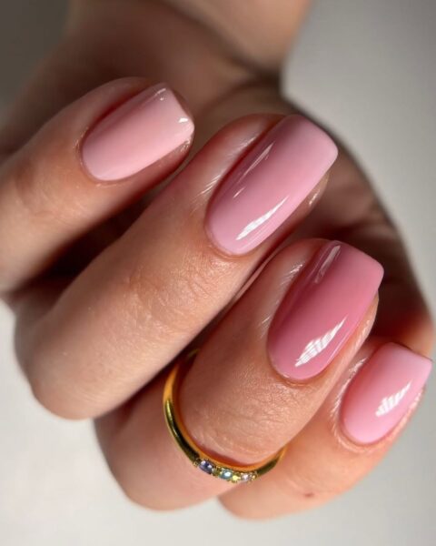 Zdjęcie dłoni ze zbliżeniem na długie, kwadratowe paznokcie pomalowane na kolor pink nude. Na środkowym palcu znajduje się złoty pierścionek. Dłoń pozuje na białym blacie.