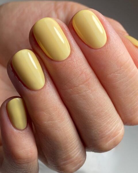 Zdjęcie dłoni ze zbliżeniem na paznokcie o krótkim, owalnym kształcie pomalowane na kolor maślanej żółci, czyli butter yellow. Dłoń pozuje na białym blacie.