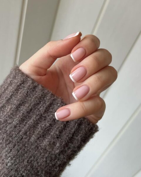 Zdjęcie dłoni ze zbliżeniem na długie, kwadratowe paznokcie ze stylizacją french manicure. Dłoń pozuje na tle białej boazerii. Widać fragment rękawa brązowego swetra.