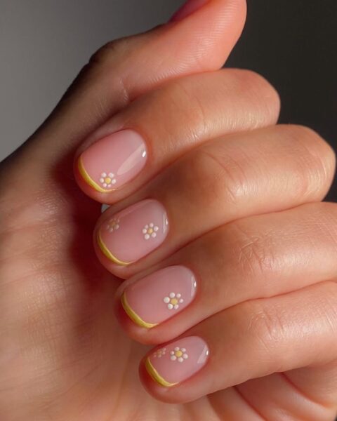 Zdjęcie dłoni ze zbliżeniem na krótkie, owalne paznokcie na koniec kwietnia w stylu french manicure z żółtą końcówką oraz drobnym wzorem kwiatów w kolorze białym.