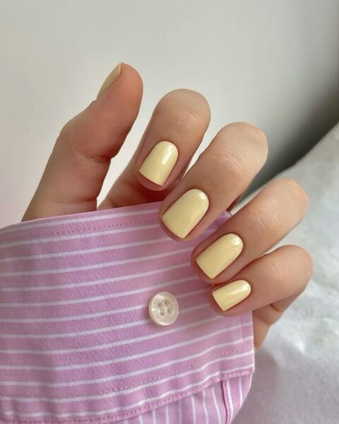 Zdjęcie dłoni ze zbliżeniem na kwadratowe paznokcie w kolorze butter yellow, czyli maślanej żółci. Widać fragment rękawa koszuli w biało-różowe paski.