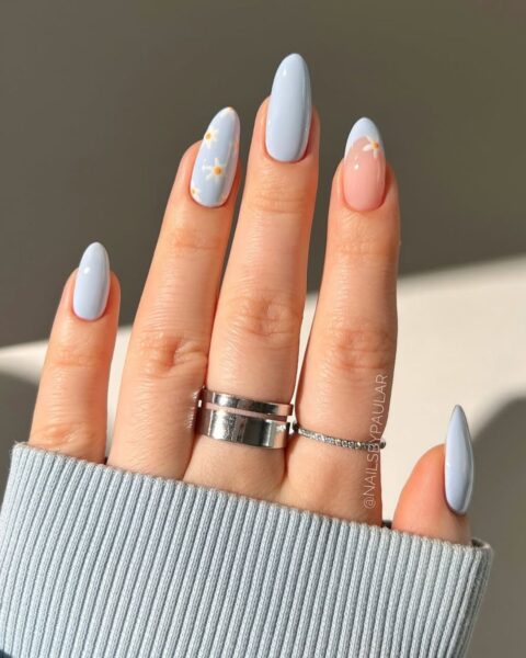 Zdjęcie dłoni z długimi, migdałowymi paznokciami w kolorze jasnego błękitu, ozdobione wzorem stokrotek, a paznokieć palca wskazującego widać french manicure z niebieską końcówką. Na palcach znajdują się liczne pierścionki w kolorze srebrnym.