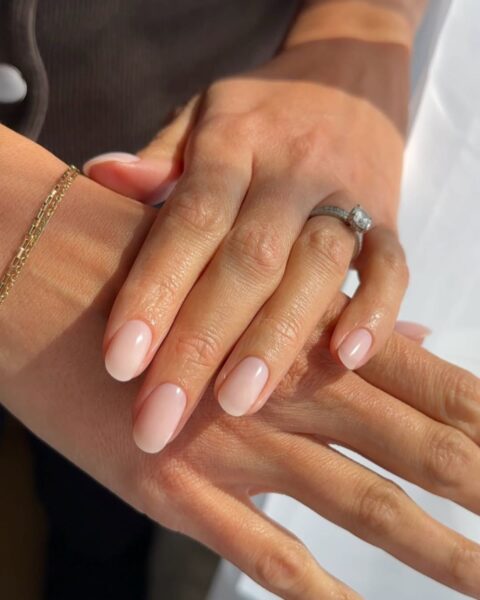 Zdjęcie dłoni ze zbliżeniem na manicure dla paznokci średniej długości. Mają owalny kształt i są pomalowane na kolor nude. Na palcu widać srebrny pierścionek z brylantem, a także złotą bransoletkę na nadgarstku.