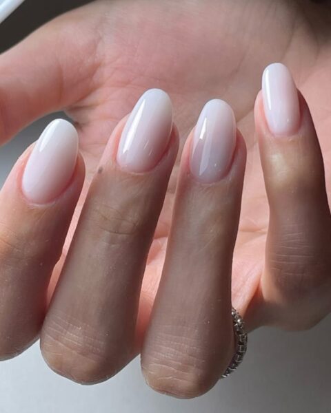 Zdjęcie dłoni z długimi, owalnymi paznokciami pomalowanymi w stylu milky nails, czyli mleczne paznokcie. Na palcu serdecznym widać pierścionek w kolorze srebrnym. Dłoń pozuje na tle białego blatu.