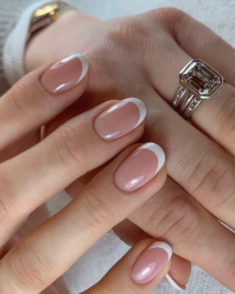 Zdjęcie dłoni z długimi, owalnymi paznokciami pomalowanymi w stylu french manicure. To zaręczynowy manicure. Dłonie układają się tak, by prezentować paznokcie oraz pierścionek w kolorze srebrnym z kamieniem na palcu serdecznym.