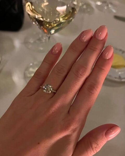 Zdjęcie dłoni ze zbliżeniem manicure zaręczynowy. Widać paznokcie w kolorze nude o owalnym kształcie. Na palcu serdecznym znajduje się srebrny pierścionek zaręczynowy z brylantem. Dłoń pozuje na tle stołu w restauracji, nakryty białym obrusem, widać w tle kieliszek, widelce i elementy zastawy.