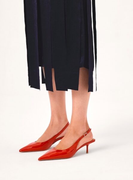 Kobiece stopy w czerwonych, lakierowanych czółenkach typu slingback z odkrytą piętą i cienkim paskiem z klamerką. Buty mają spiczasty nosek i niski obcas typu kitten heel. Stylizacja uzupełniona jest granatową spódnicą z frędzlami sięgającą połowy łydki.