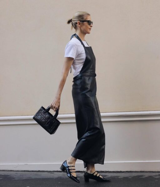 Kobieta idąca ulicą w eleganckim, minimalistycznym stroju. Ma na sobie długą, czarną sukienkę na szelkach wykonaną z ekoskóry, założoną na biały t-shirt. Niesie czarną, pikowaną torebkę w dłoni i nosi lakierowane czarne buty z trzema paskami. Jej włosy są spięte w niski koczek, a twarz osłaniają duże okulary przeciwsłoneczne.