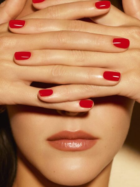 Zbliżenie twarzy kobiety z dłońmi zakrywającymi oczy, prezentującymi perfekcyjny manicure w intensywnym, klasycznym odcieniu czerwieni z kolekcji Chanel Le Vernis. Usta w naturalnym kolorze z delikatnym połyskiem.