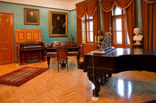 Dom pianisty Franciszka Liszta w Budapeszcie. Elegancki pokój muzealny z zabytkowymi instrumentami klawiszowymi, w tym fortepianem i organami, ozdobiony portretami i popiersiami kompozytorów; na ścianach wzorzysta tapeta, a przy oknach ciężkie zasłony w kolorze miedzianym.