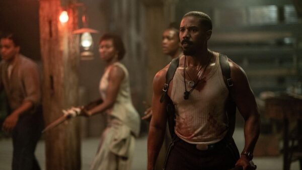 Grupa osób w napiętej scenie filmowej w ciemnym, zadymionym pomieszczeniu. Na pierwszym planie mężczyzna w zabrudzonej i zakrwawionej koszulce na ramiączkach, z napiętym wyrazem twarzy, noszący uprząż i naszyjnik. W tle trzy osoby, jedna z nich trzyma strzelbę. Scena oświetlona ciepłym, przytłumionym światłem latarni. Jest to kadr z filmu "Grzesznicy".