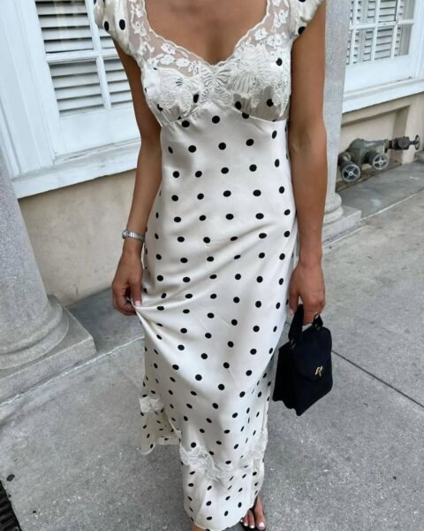 Kobieta w eleganckiej, wiosennej stylizacji na wesele pozuje na tle jasnej elewacji budynku. Ma na sobie długą, satynową sukienkę w kolorze ecru z czarnymi groszkami, ozdobioną delikatną koronką przy dekolcie i na dole. Sukienka ma kobiecy, dopasowany krój i subtelny połysk. W dłoni trzyma małą, czarną torebkę, a na nadgarstku nosi srebrny zegarek. Stylizacja jest romantyczna i idealna na wiosenne, eleganckie przyjęcie.