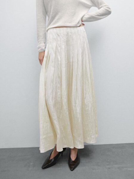 Modelka w jasnej, plisowanej spódnicy o długości maxi, połączonej z białym, długim swetrem. Do stylizacji dobrano czarne, eleganckie szpilki. Tło jest minimalistyczne, w jednolitym, pastelowym kolorze. Spódnica jest z Massimo Dutti.