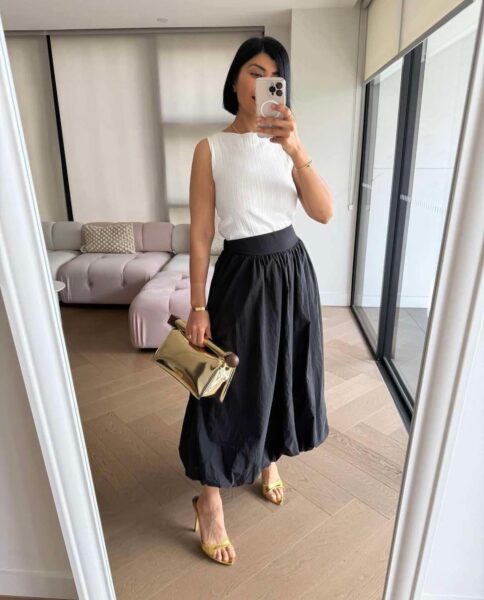 Kobieta robi selfie w lustrze, prezentując elegancką i minimalistyczną stylizację. Ma na sobie biały, bezrękawnikowy top z delikatną fakturą oraz czarną, rozkloszowaną spódnicę o długości midi. Stylizację uzupełniają złote sandałki na obcasie i połyskująca, złota torebka kopertowa z kulkowym zapięciem. Kobieta stoi w nowoczesnym, jasnym wnętrzu z dużymi oknami i stylową sofą w tle.