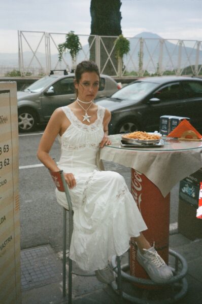 Kobieta w białej sukni siedzi przy małym stoliku na zewnątrz włoskiej restauracji ulicznej, z talerzem spaghetti przed sobą. Ma na sobie białe sportowe buty i dużą naszyjnik w kształcie gwiazdy. W tle widać zaparkowane samochody i ogrodzenie z widokiem na górzysty krajobraz. Jest do zdjęcie z nowej kampanii INUIKII na wiosnę i lato 2025.