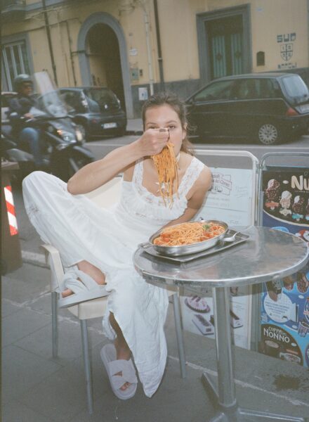 Kobieta w białej sukni siedzi przy metalowym stoliku ulicznej restauracji, energicznie jedząc spaghetti z dużego talerza. Na stopach ma jasne klapki, a w tle widać przejeżdżające skutery, zaparkowane samochody i reklamę lodów. Jest to zdjęcie z kampanii INUIKII na wiosnę i lato 2025.