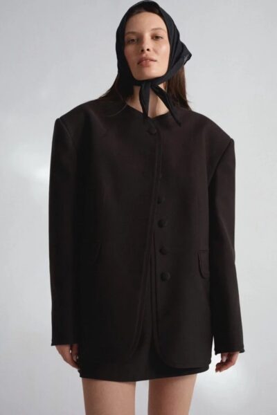 Modelka ubrana w oversize’owy czarny płaszcz z dużymi guzikami i czarną chustką zawiązaną pod brodą, na tle neutralnego tła.