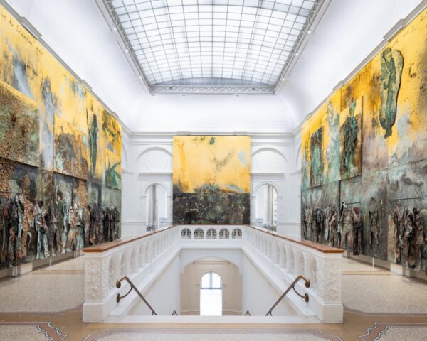 Wnętrze eleganckiej sali muzealnej z wysokim, przeszklonym sufitem. Na ścianach wiszą monumentalne, ekspresyjne obrazy o żółto-brązowej kolorystyce, autorstwa Anselma Kiefera. Dolne części dzieł mają trójwymiarowe elementy przypominające ubrania lub figury, co nadaje pracom rzeźbiarski charakter.