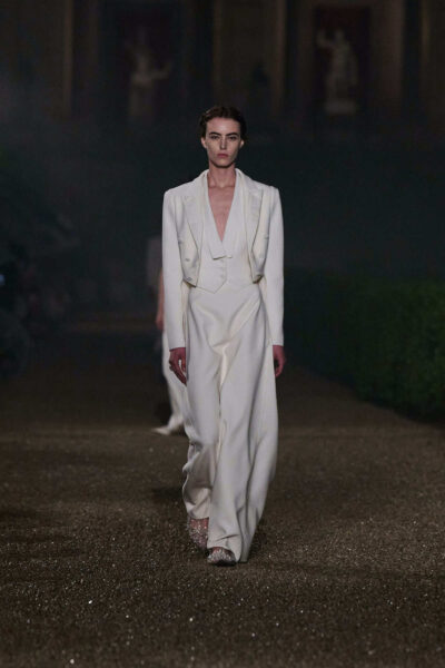 Modelka w eleganckim, białym smokingu haute couture z krótką marynarką z dwurzędowymi guzikami, głębokim dekoltem i szerokimi spodniami, idzie po żwirowym wybiegu nocą.