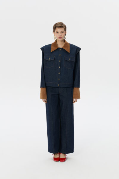Studyjne zdjęcie lookbookowe: modelka stoi frontalnie na białym tle w oversize’owym komplecie z ciemnego denimu – kurtce z szerokimi ramionami, dużym brązowym kołnierzem i mankietami oraz w szerokich spodniach; ma czerwone lakierowane baleriny i złote kolczyki-koła. Letnia kolekcja polskiej marki Naji.