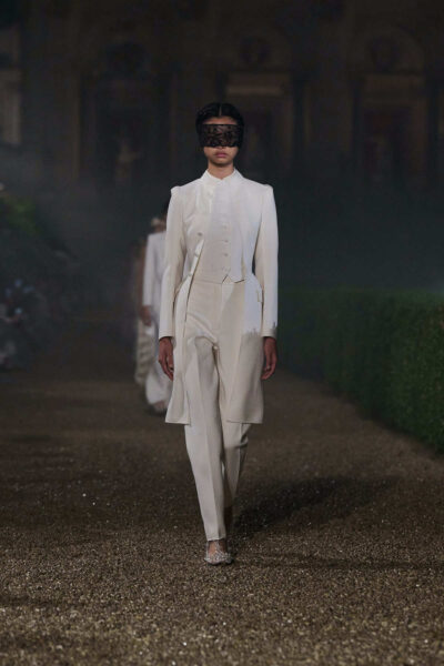 Modelka w białym, trzyczęściowym garniturze haute couture z długą marynarką frakową, kamizelką i koronkową maską, uosabiająca androgyniczną elegancję.