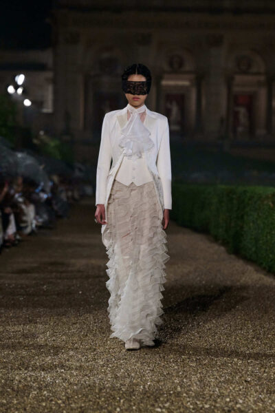 Modelka w białym żakiecie haute couture z falbaną na piersi, zestawionym z długą spódnicą z wielu warstw tiulu, nosi czarną, koronkową maskę zasłaniającą oczy, tworząc teatralny i romantyczny efekt.