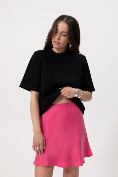 Modelka na białym tle w minimalistycznym zestawie: czarny, luźny T-shirt z krótkim rękawem i różowa, satynowa minispódniczka z lekkim połyskiem. Stylizację uzupełniają masywne srebrne pierścionki i bransoletka.