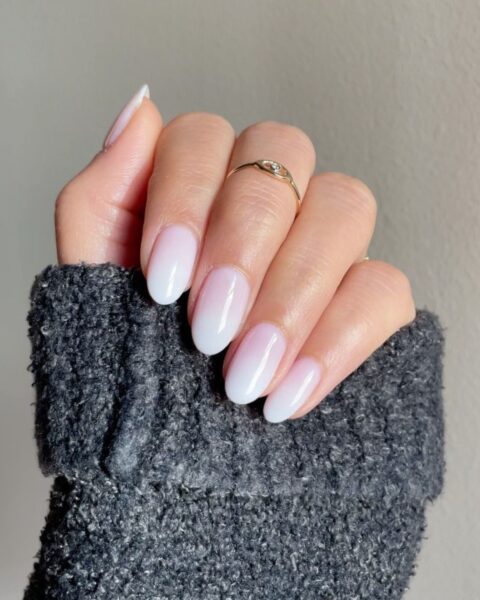 Zdjęcie dłoni ze zbliżeniem na długie, owalne paznokcie ze wzorem french ombré manicure. Na palcu środkowym znajduje się złoty pierścionek. Widać fragment szarego rękawa swetra.